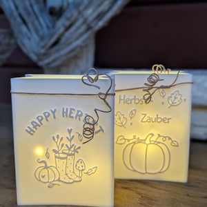 Könnte beinhalten: Zwei beleuchtete, weiße, rechteckige Laternen mit dekorativen Ausschnitten und Text. Auf einer steht "HAPPY HER" mit einem Kürbis, Stiefeln und einem Pilz-Design. Die andere sagt "Herbst Zauber" mit einem Kürbis und Blättern.