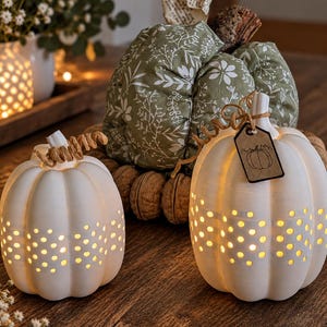 May include: Two white ceramic pumpkins with perforated dot patterns and internal warm lighting, featuring brown curly stems and a small rectangular tag with a pumpkin drawing. Behind them sits a fabric pumpkin in sage green with white botanical prints.
