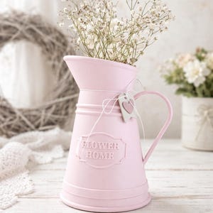Könnte beinhalten: Eine rosa Metallkanne mit Henkel, gefüllt mit weißen Blumen. Auf der Kanne steht der Schriftzug "FLOWER HOME". Ein kleines herzförmiges Etikett hängt am Henkel. Die Kanne steht auf einer weißen Holzoberfläche.
