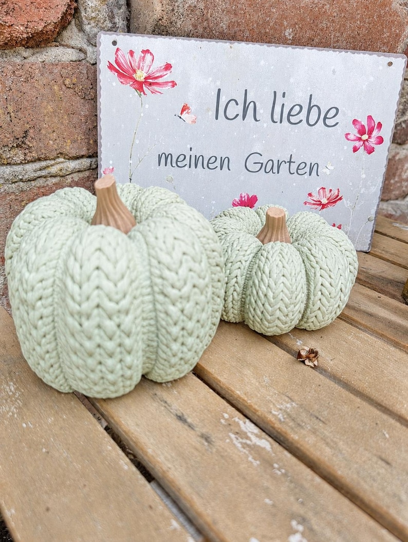May include: Two sage green pumpkins with a textured knit pattern and brown stems. Behind them is a rectangular metal sign with a light gray background, red flower illustrations, and the German text Ich liebe meinen Garten.