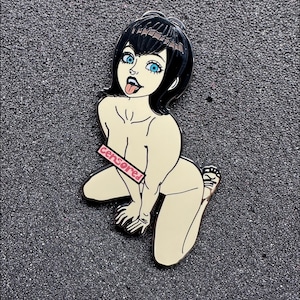 May include: Enamel pin depicting a cartoon woman with black hair and blue eyes, kneeling with her tongue out. The word "Censored" is printed on a pink rectangle. The pin has a black outline and a glossy finish.