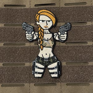 May include: Embroidered patch depicting a cartoon woman with orange hair in a plait, holding two pistols. She wears a camouflage top and shorts with white straps. The patch is set against a brown, textured background.