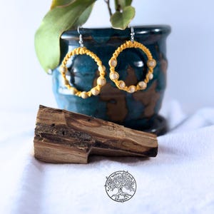 May include: A pair of handmade hoop earrings. The earrings feature a yellow woven design with small, light-colored beads. The earrings are displayed in front of a blue and brown ceramic pot with a green plant.