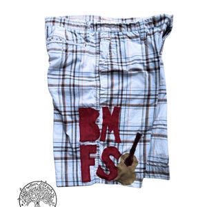 May include: Light blue plaid shorts featuring a dark red corduroy applique of the letters BMFS and a brown corduroy guitar shape. The shorts have a waistband with belt loops and a classic button and zipper fly closure on the front.