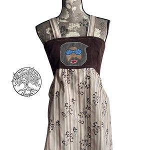 May include: A brown corduroy apron with a striped skirt. The apron features a patch with a cartoon face wearing blue sunglasses and a mustache. The apron has adjustable straps and is displayed on a mannequin.