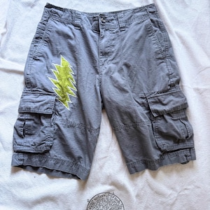 May include: Grey cargo shorts featuring a lime green lightning bolt patch on the right leg, a button and zipper fly, two side cargo pockets with flaps, and a circular logo at the bottom with the text Seekers Finds Designs.