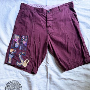 May include: Maroon shorts featuring a button closure and a waistband with belt loops. The right leg displays a large, multi-colored fabric appliqué patch with the letters BMFS and a small acoustic guitar graphic stitched onto the fabric surface.