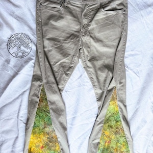 May include: Tan-colored jeans with a unique design. The jeans feature a colorful, patterned fabric insert on the outer leg, showcasing shades of green, yellow, and red. The jeans are laid flat on a white surface.