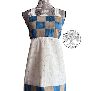 May include: An apron with a corduroy checkered top in blue and tan, and a white floral patterned bottom. The apron has white straps and a logo with the text "Seekers Finds Designs".