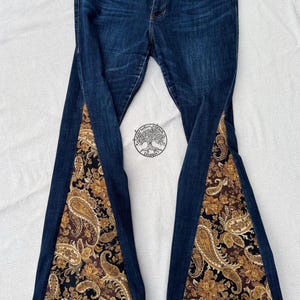 May include: Dark blue denim jeans featuring wide, triangular inserts at the lower legs. The inserts are made of a black fabric with a detailed gold and brown paisley floral pattern. The jeans have a standard button closure and front pockets.