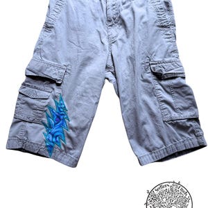May include: Light gray cargo shorts featuring a button closure, belt loops, and two side cargo pockets. A blue and teal lightning bolt patch with a swirling pattern is stitched onto the lower right leg. A circular logo with a tree and the text Seekers Finds Designs is visible in the corner.