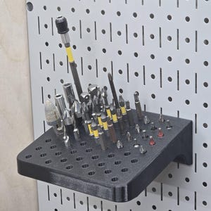 May include: A black 3D-printed pegboard shelf filled with various drill bits and driver bits. The shelf is mounted on a white pegboard. The bits are a mix of silver, black, and yellow. The shelf has multiple holes to hold the bits.
