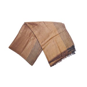 May include: A rectangular scarf in shades of brown and gold featuring a central diamond pattern. The scarf includes sections with vertical stripes and a fringed edge on one side, showcasing a woven texture throughout the entire fabric surface.