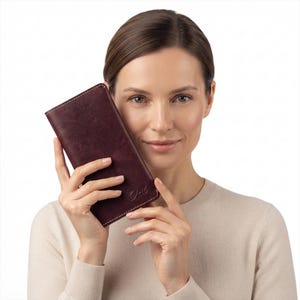 May include: A rectangular dark burgundy leather wallet featuring visible white stitching along the edges and a debossed logo on the bottom right corner. The wallet has a smooth, matte texture and a slim, vertical orientation with rounded corners.