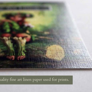 May include: A close-up of a printed image on high-quality fine art linen paper. The image features a whimsical fairy-like figure with green skin and long, flowing hair. The fairy is sitting on a mossy rock in a lush forest setting.