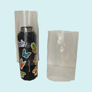 Puede incluir: Una botella de agua de acero inoxidable negra con una variedad de pegatinas coloridas, incluyendo una mariposa, una piña y un hongo. La botella está dentro de una bolsa de plástico transparente.