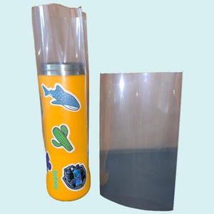 Puede incluir: Un vaso amarillo con varias pegatinas, incluyendo un tiburón, un cactus y una flor. El vaso está envuelto en una envoltura retráctil de plástico transparente.