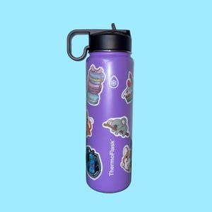 Puede incluir: Una botella de agua ThermoFlask morada con tapa y asa negras. La botella está decorada con pegatinas coloridas, incluyendo macarons, una magdalena, un koala y un corgi. El fondo es azul claro.