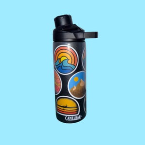 Puede incluir: Botella de agua CamelBak negra con tapa y asa negras. La botella está cubierta con pegatinas circulares de colores que representan paisajes, incluyendo montañas, puestas de sol y olas del océano. El logotipo de CamelBak está impreso en la parte inferior.