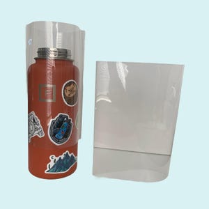 Puede incluir: Una botella de agua naranja brillante con una variedad de pegatinas, incluyendo una cordillera, un oso, un erizo y un logotipo. La botella está envuelta en plástico transparente.