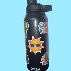 Puede incluir: Botella de agua Camelbak negra con tapa y asa negras. La botella está decorada con pegatinas coloridas, incluyendo un cactus, un sol con gafas de sol, un elefante y las palabras "Good Vibes."