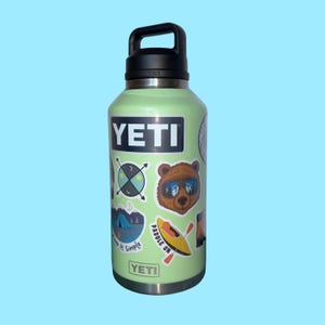 Puede incluir: Una botella de agua YETI verde claro con asa y tapa negras. La botella tiene varias pegatinas, incluyendo un oso con gafas de sol, un kayak y las palabras "Keep It Simple". La botella mide aproximadamente 25 cm de alto.