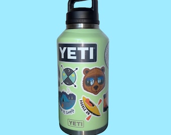 May include: A light green YETI water bottle with a black handle and lid. The bottle has several stickers, including a bear wearing sunglasses, a kayak, and the words "Keep It Simple". The bottle is approximately 10 inches tall.
