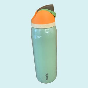 Puede incluir: Una botella de agua aislada de color turquesa con una tapa naranja y una banda blanca. La tapa tiene un acento gris y verde. La marca "owala" está impresa en la botella. La botella está diseñada para mantener las bebidas frías durante horas.