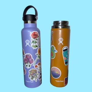 Puede incluir: Dos botellas de agua Hydro Flask. Una es lavanda con pegatinas florales, un asa negra y una tapa plateada. La otra es naranja con pegatinas de aguacate y paisaje. Ambas botellas tienen el logotipo de Hydro Flask.