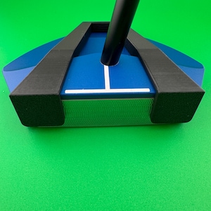 Könnte beinhalten: Ein blauer und schwarzer Golfschläger mit schwarzem Schaft. Der Schlägerkopf hat eine blaue Oberfläche mit einer weißen Ausrichtungslinie. Die Kanten des Schlägerkopfes sind schwarz. Der Schläger liegt vor einem grünen Hintergrund.
