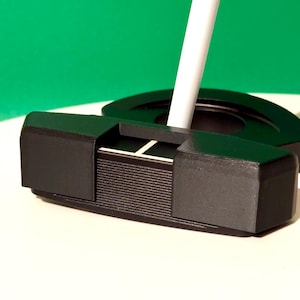 Puede incluir: Un putter de golf negro con un eje blanco, sobre un fondo verde. La cabeza del putter tiene un diseño rectangular con líneas horizontales y una línea blanca para la alineación. Este equipo está diseñado para la precisión y el control en el green.