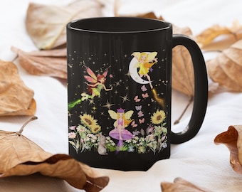 May include: A black ceramic mug featuring a colorful graphic of three fairies, a crescent moon, sunflowers, daisies, a small rabbit, and two frogs. The design includes golden star accents and small butterflies scattered across the dark background.