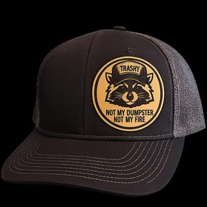 May include: A black trucker hat with a gray mesh back. The front features a circular patch with a raccoon wearing a hat and the text "TRASHY". Below the raccoon is the text "NOT MY DUMPSTER NOT MY FIRE".