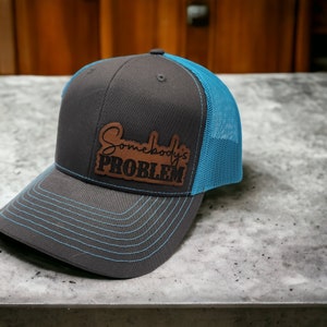 May include: A gray trucker hat with a blue mesh back and brim. The hat features the text "Somebody's PROBLEM" on a brown leather patch. The brim has blue stitching.