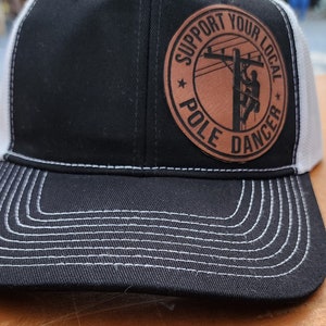 May include: Black and white trucker hat with a brown leather patch. The patch features the text "SUPPORT YOUR LOCAL POLE DANCER" around an image of a lineman on a utility pole. The hat has a curved brim and white mesh back.