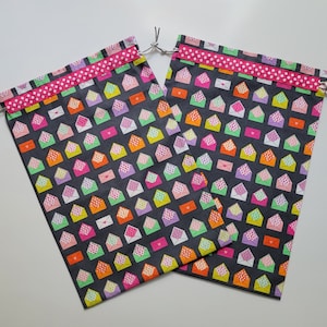 May include: Two drawstring bags with a dark gray background covered in colorful envelope patterns. Each envelope is a different color, including pink, orange, green, and purple. A pink polka dot ribbon trims the top, and a gray drawstring is visible.