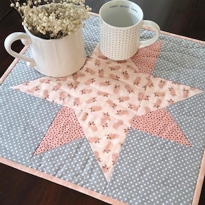 May include: A gray and pink star-shaped quilted table runner with a white mug that says "home sweet home" on it. The mug is sitting on the table runner.
