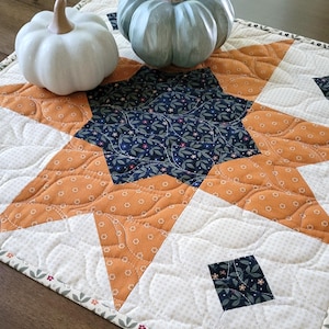 May include: A quilted table runner with a starburst design in orange, navy, and white, adorned with two decorative pumpkins. The runner features a floral pattern and is placed on a wooden surface.