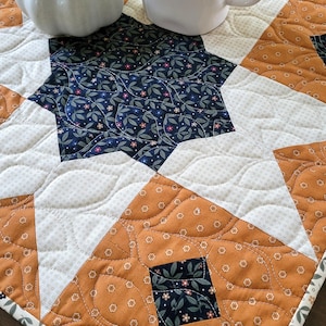 May include: A quilted table runner with a star pattern in cream, orange, and navy blue floral fabric. A white ceramic mug with the word "LOVELY" and a small white pumpkin are on the runner.