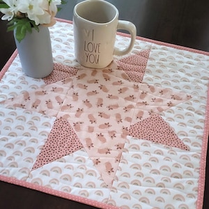 May include: A pink and white quilted table runner with a star design. The runner is made of different fabrics with various patterns, including pink pigs, white polka dots, and pink and white rainbows. The runner is trimmed with a pink and white polka dot border. A white mug with the text "I Love You" is sitting on the runner.