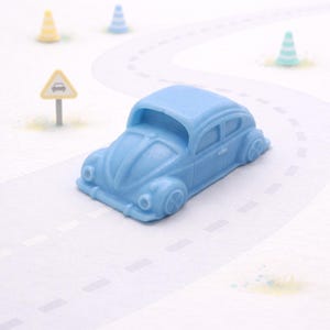 Puede incluir: Un coche de juguete azul claro se encuentra sobre una carretera blanca con líneas discontinuas. El coche tiene un diseño clásico y redondeado. También son visibles pequeños conos de tráfico y una señal de tráfico.