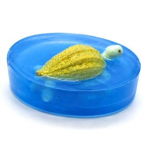 Pode incluir: Um sabonete azul translúcido de formato oval com uma pequena figura de tartaruga amarela texturizada incrustada no centro. A tartaruga tem uma cabeça clara e um casco detalhado e irregular, parecendo que está a nadar dentro do sabonete.