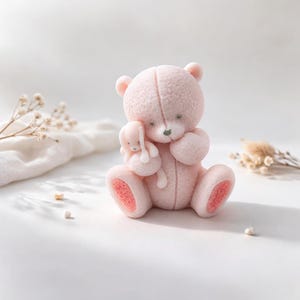 以下が含まれることがあります： 小さなウサギの置物を抱いた、薄いピンク色のテディベアの置物。クマは質感のある表面で、足と耳にピンクのアクセントがあります。ウサギもピンク色です。背景は白です。