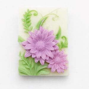 May include: A rectangular soap bar with a floral design. It features two purple flowers and green leaves on a cream background. The flowers are the focal point, with detailed petals and a textured surface.