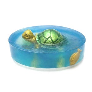 Pode incluir: Uma barra de sabão redonda e azul com um desenho de tartaruga verde e dourada. A tartaruga parece estar nadando na água. Um peixe dourado também é visível no sabão.