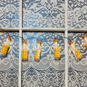 May include: String of decorative lights featuring miniature liquor bottle-shaped bulbs. Each clear plastic bottle has an orange cap and a yellow label with the text "99 PEACHES". The string is against a white lace curtain.