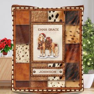 May include: A rectangular throw blanket featuring a patchwork design of brown and white animal fur patterns. The center displays a horse wearing a Santa hat and scarf with gifts, labeled EMMA GRACE and JOHNSON in a brown frame with stitching.