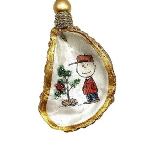 May include: A gold-painted oyster shell ornament with a hand-painted image of Charlie Brown holding a small Christmas tree. The ornament has a red and gold hanging loop.