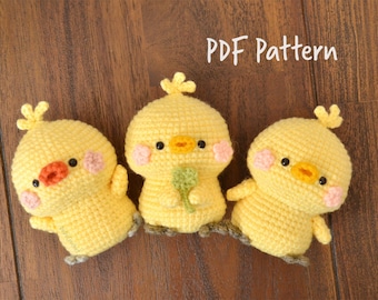 May include: Three crocheted yellow chick amigurumi toys with black eyes, orange beaks, and pink cheeks. The middle chick is holding a green leaf. The text "PDF Pattern" is written in white on a brown wooden background.