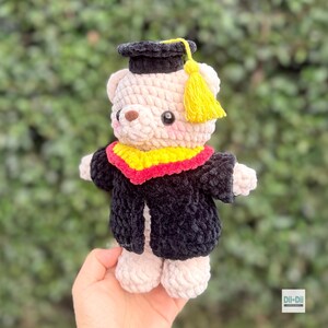 Peut inclure: Un ours en peluche crochet&eacute; portant une tenue de remise de dipl&ocirc;me. L'ours est de couleur cr&egrave;me avec une toque et une robe noires. Le col de remise de dipl&ocirc;me est jaune et rouge. L'ours a un nez brun et des yeux noirs. Une gland jaune pend de la toque.